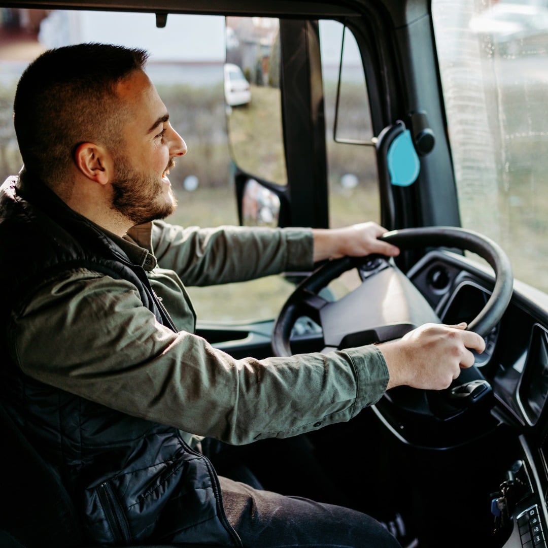 A smiling male driver in a black vest and green shirt, sitting behind the wheel and driving a truck.