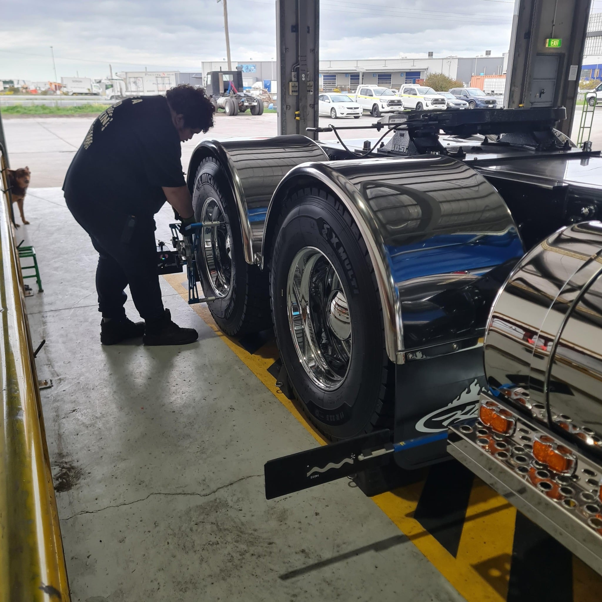A mechanic in a workshop performs a wheel alignment on the rear wheels of a large semi-truck chassis.