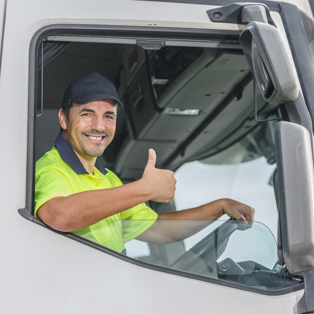 A smiling male truck driver in a neon yellow polo shirt and dark cap, sitting in the cab of a white truck and giving a thumbs-up.