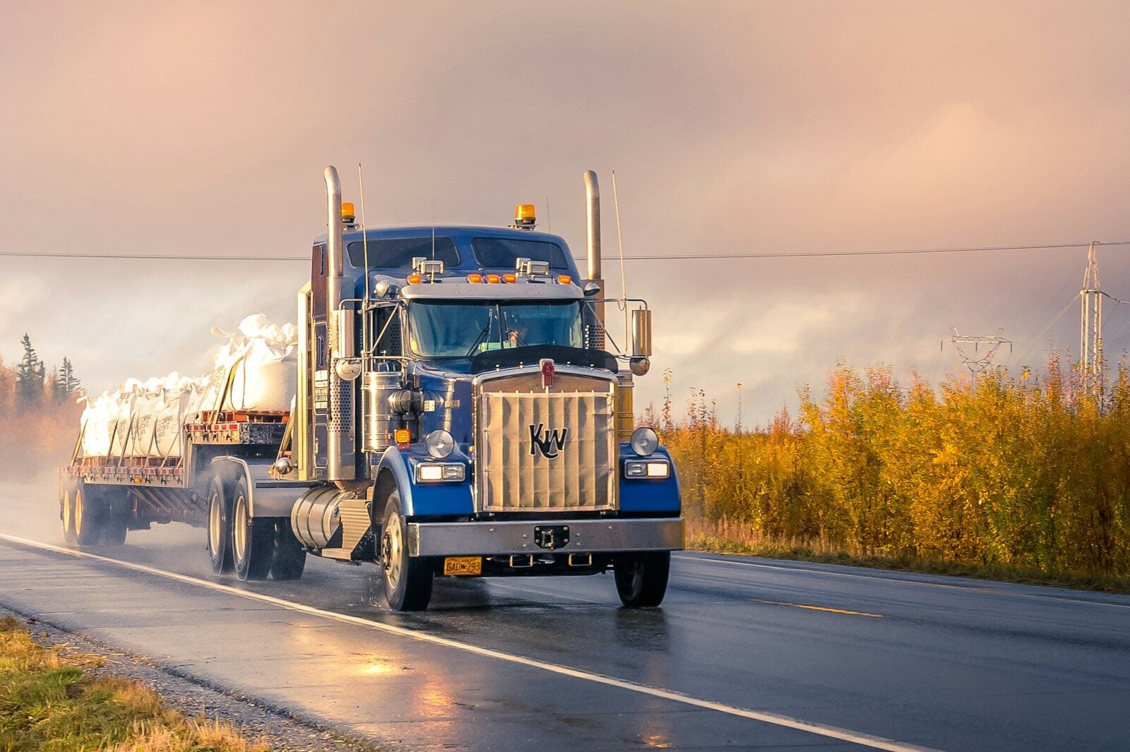 A blue Kenworth semi-truck pulling a flatbed trailer loaded with large white bulk bags