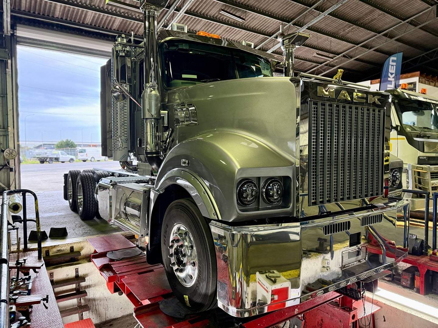 A shiny, silver Mack truck is positioned over a red service pit inside a garage.
